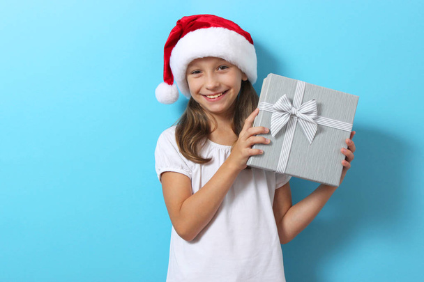 linda chica alegre en un sombrero de Navidad en un fondo coloreado sosteniendo un regalo. - Foto, imagen