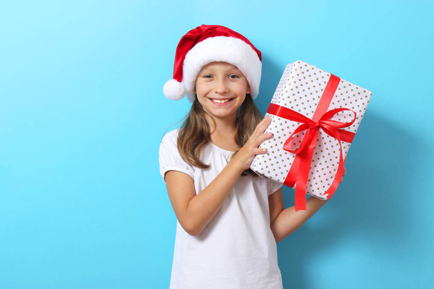 cute cheerful girl in a Christmas hat on a colored background holding a gift - Photo, Image