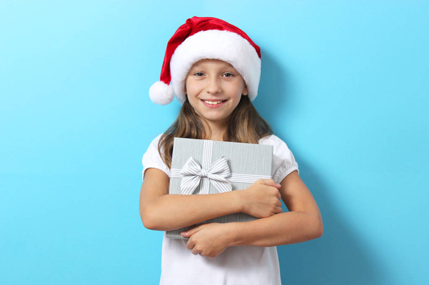 χαριτωμένο χαρούμενο κορίτσι σε ένα καπέλο Χριστουγέννων σε ένα έγχρωμο φόντο κρατώντας ένα δώρο - Φωτογραφία, εικόνα