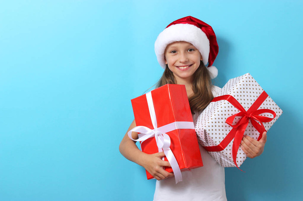 schattig vrolijk meisje in een kerst hoed op een gekleurde achtergrond met geschenken in haar handen - Foto, afbeelding