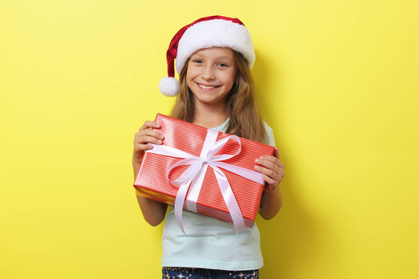 linda chica alegre en un sombrero de Navidad en un fondo coloreado sosteniendo un regalo. - Foto, Imagen