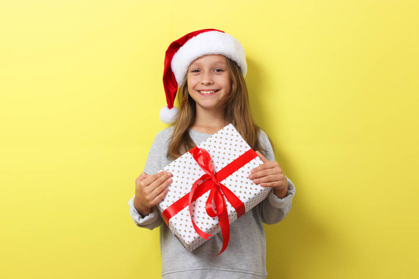 niedliche fröhliche Mädchen in einem Weihnachtsmütze auf einem farbigen Hintergrund hält ein Geschenk - Foto, Bild