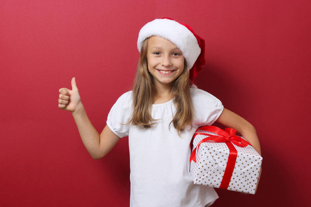 cute cheerful girl in a Christmas hat on a colored background holding a gift - Valokuva, kuva
