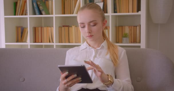 Closeup portrait of young pretty caucasian blonde female student using the tablet looking at camera smiling cheerfully sitting on the couch indoors in the apartment - Materiaali, video