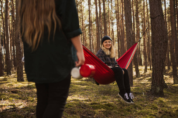 Happy young hipster girl enjoys life and nature on hammock with other woman in the summer forest. - Фото, изображение