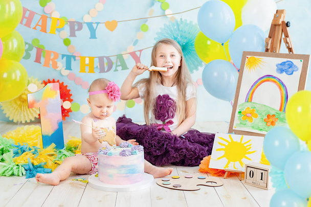 Dekoration für den ersten Geburtstag eines Mädchens, Kuchen im Stil eines Kunstmalers zertrümmern. Baby lächelt in die Kamera. Porträt eines entzückenden kaukasischen Jungen, der auf Holzboden im Atelier sitzt. - Foto, Bild