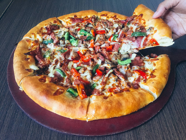 mano humana tomando rebanada de pizza con carne y verduras servidas en mesa oscura
 - Foto, Imagen