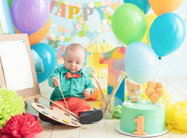 decoration for boy's first birthday, smash cake in a art painter style. baby smiling looking at camera. Portrait of adorable caucasian baby boy celebratingsitting on wooden floor in studio. - Photo, image