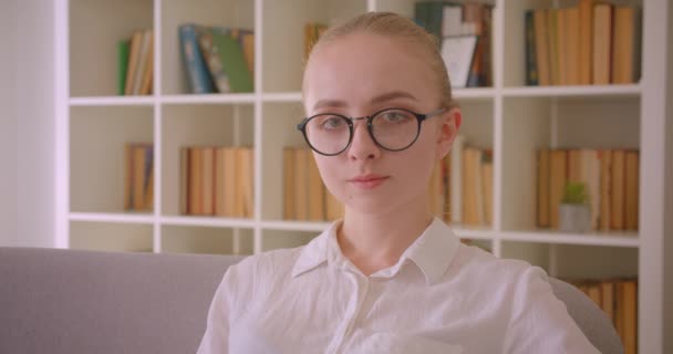 Closeup portrait of young pretty caucasian blonde female student in glasses looking at camera smiling sitting on the sofa indoors in the apartment - Video