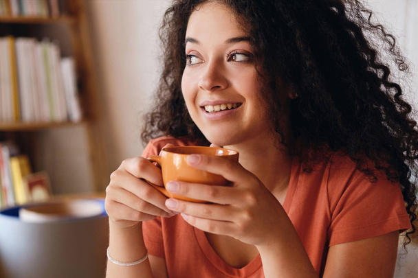 Πορτρέτο της νεαρής ελκυστική χαμογελαστή γυναίκα με σκούρα σγουρά μαλλιά κρατώντας πορτοκαλί κούπα καφέ στα χέρια καλά κοιτάζοντας στην άκρη στο άνετο σπίτι μόνος - Φωτογραφία, εικόνα