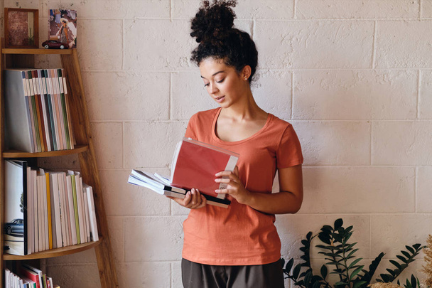 T-shirt koyu kıvırcık saçlı genç muhteşem kadın rahat evde kitaplık yakınında duran ellerde kitap tutan - Fotoğraf, Görsel