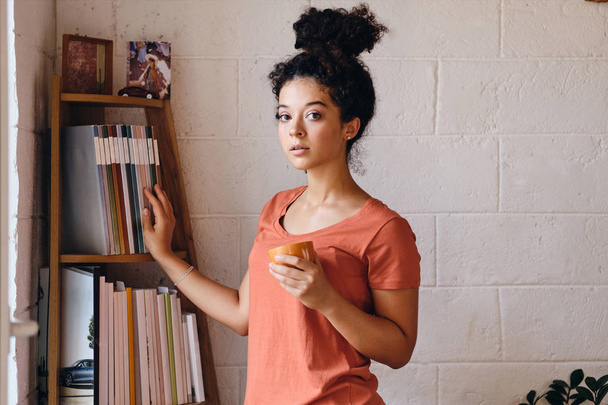 Mujer atractiva joven con pelo rizado oscuro en camiseta sosteniendo taza de café en la mano cuidadosamente mirando en la cámara de pie cerca de la estantería en casa acogedora
 - Foto, imagen