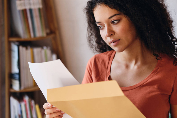 Jovem mulher séria com cabelos encaracolados escuros em T-shirt olhando atenciosamente na carta com resultados do exame em mãos em casa aconchegante
 - Foto, Imagem