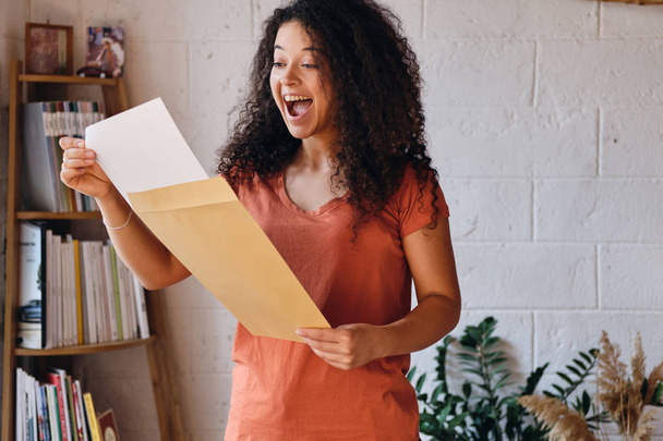 Jonge vrolijke vrouw met donkere krullend haar in T-shirt gelukkig opening envelop met examen resultaten brief bij Cozy Home - Foto, afbeelding