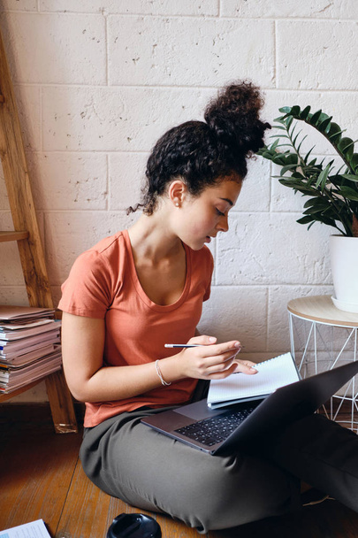 Mladá studentka s tmavými kudrnatými vlasy, sedící na podlaze s laptopem na kolenou zamyšleně studující v útulném domě - Fotografie, Obrázek