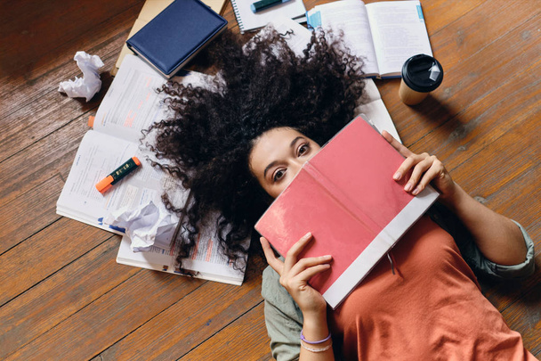 Young pretty student girl with dark curly hair lying on floor with textbooks around dreamily looking in camera covering mouth with notebook at home - Foto, Imagem