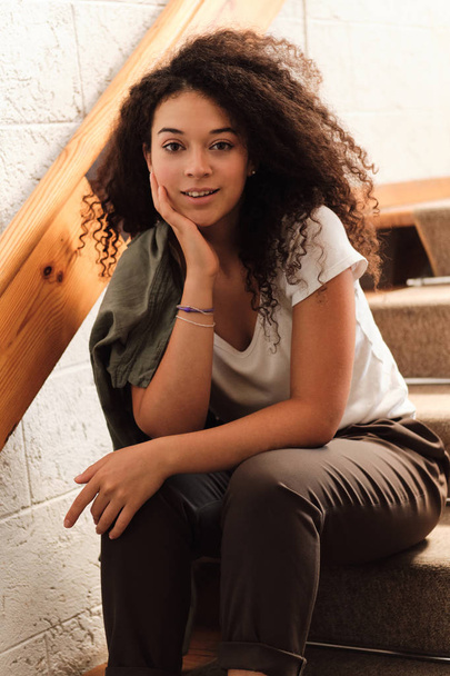 Young beautiful smiling woman with dark curly hair sitting on stairs in university leaning head on hand and dreamily looking in camera - Foto, immagini