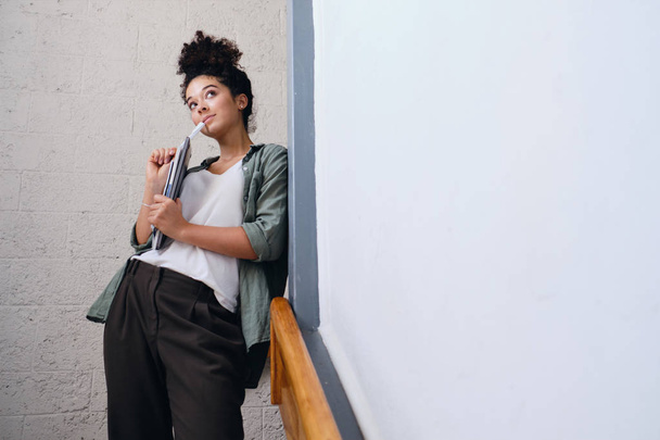 Young beautiful student girl with dark curly hair in khaki shirt and pants leaning on wall in university with textbooks in hands and dreamily looking aside - Foto, Bild