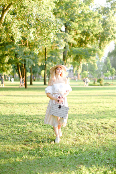 Young pretty american woman in hat walking in park and keeping bag and sunglasses. - Φωτογραφία, εικόνα