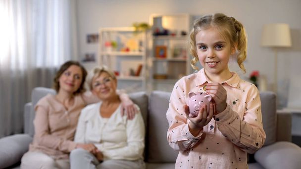 Schoolgirl putting coin in piggybank, proud mother and granny behind, savings - Zdjęcie, obraz
