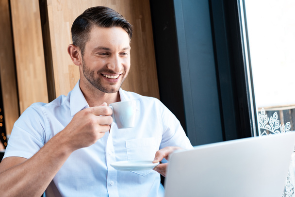 smiling freelancer holding saucer and cup of coffee while looking at laptop sceeen - Foto, afbeelding