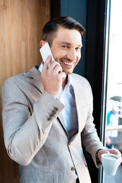 smiling businessman holding cup of coffee and talking on smartphone in cafe - Фото, зображення