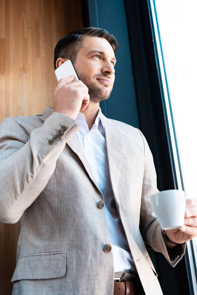 smiling businessman holding cup of coffee and talking on smartphone in cafe - Foto, imagen