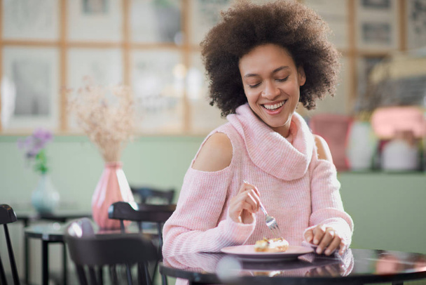 Porträt einer charmanten positiven Mischlingsfrau im rosafarbenen Rollkragenpullover, die in einer Konditorei sitzt und Torte isst. - Foto, Bild