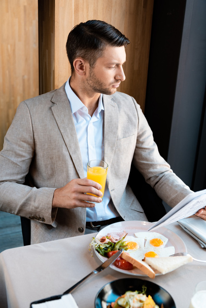 pensive businessman holding glass of orange juice and newspaper in cafe - Photo, Image