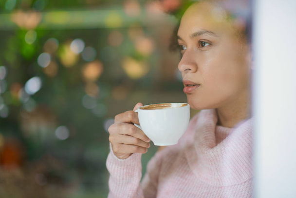 Superbe femme mixte en pull col roulé rose debout dans un café, buvant de l'espresso et regardant par la fenêtre
. - Photo, image