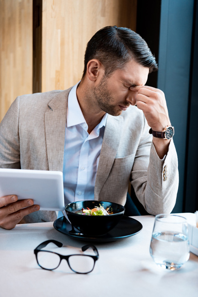 hombre de negocios cansado tocando la nariz con los ojos cerrados y sosteniendo la tableta digital en la cafetería
 - Foto, Imagen