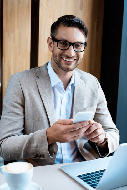 smiling businessman in glasses holding smartphone while sitting at table with laptop in cafe - Fotó, kép