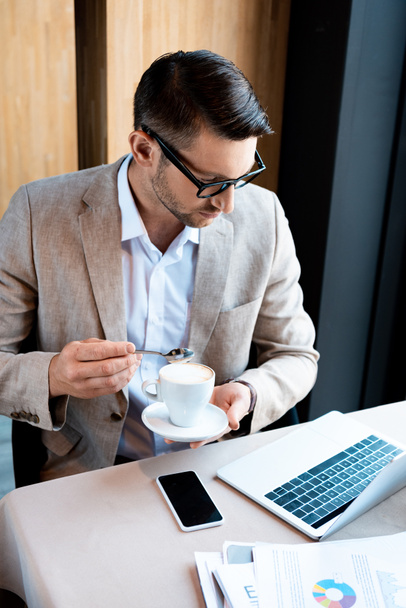 focused businessman in glasses holding cup of coffee while sitting at table with laptop in cafe - Fotoğraf, Görsel