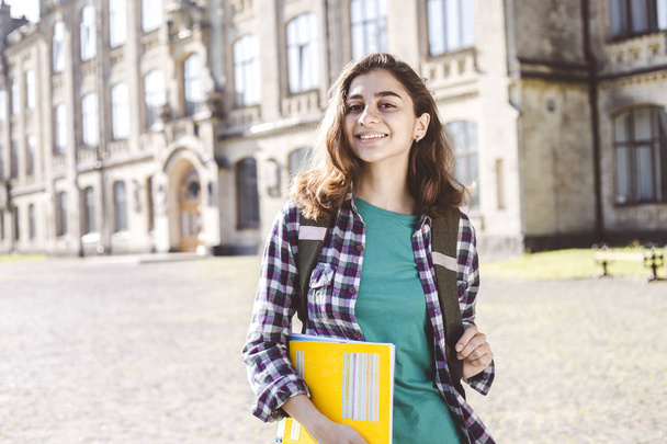 笑顔のインドの若い女子学生は、大学の背景に立って教育書を保持しています.大学の近くに幸せなブルネットの女の子.  - 写真・画像