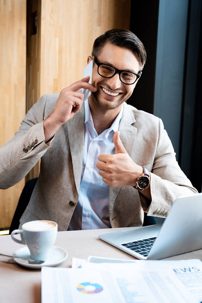 hombre de negocios sonriente en gafas hablando en smartphone y mostrando el pulgar hacia arriba en la cafetería
 - Foto, imagen