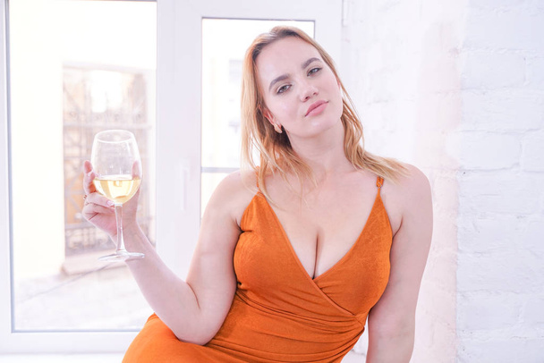 自宅の窓ガラスにオレンジ色のドレスに座り、大きなグラスからアルコール白ワインを飲むプラスサイズのフィギュアを持つ美しい女の子 - 写真・画像