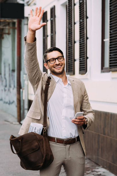 usmívající se obchodník v brýlích, který drží smartphone a mával rukou na ulici - Fotografie, Obrázek