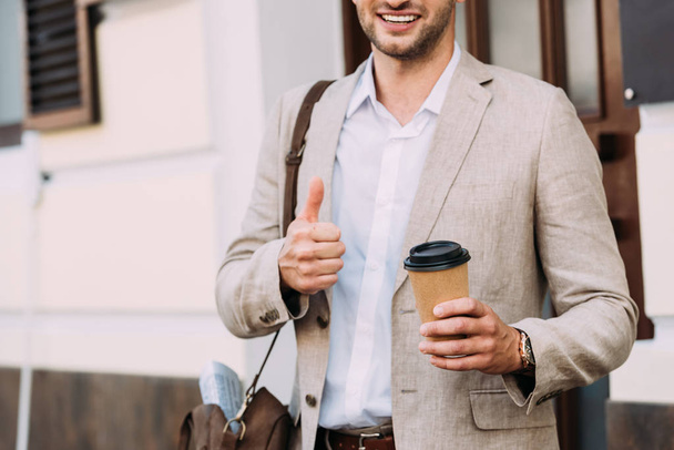 recortado vista de sonriente hombre de negocios en vasos sosteniendo café para ir y mostrando el pulgar hacia arriba en la calle
 - Foto, Imagen
