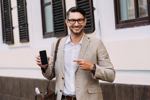front view of smiling businessman pointing with finger at smartphone with blank screen on street - Фото, изображение