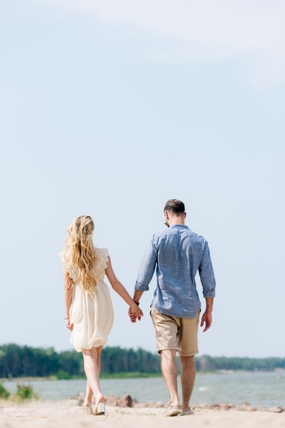 back view of adult couple walking along beach and holding hands at sunny day - Фото, изображение