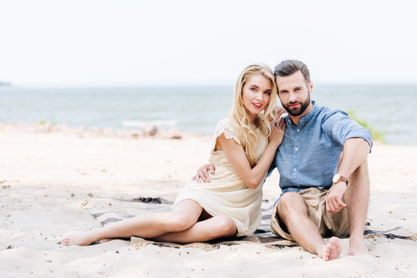 beautiful young barefoot couple sitting on blanket at beach near sea - Foto, imagen