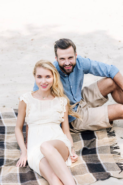 beautiful smiling young couple sitting on plaid blanket at sandy beach - Fotografie, Obrázek