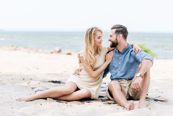 happy beautiful young barefoot couple sitting on blanket and looking at each other at beach near sea - 写真・画像