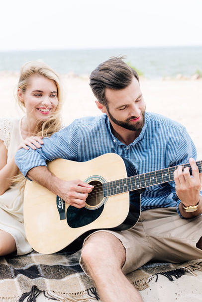 smiling beautiful young barefoot woman sitting on blanket near boyfriend with acoustic guitar at beach near sea - Фото, изображение