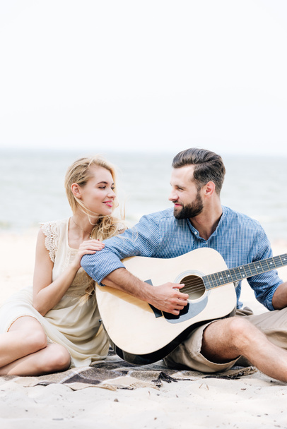smiling beautiful young barefoot woman sitting on blanket and looking at boyfriend with acoustic guitar at beach near sea - Foto, Imagen