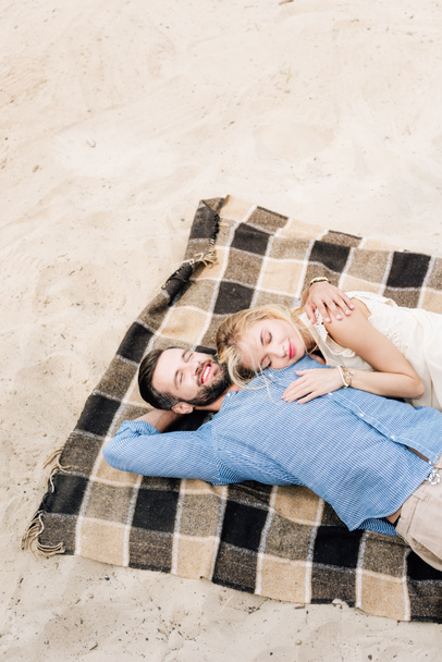 happy couple lying together with closed eyes on checkered blanket at sandy beach - Photo, Image