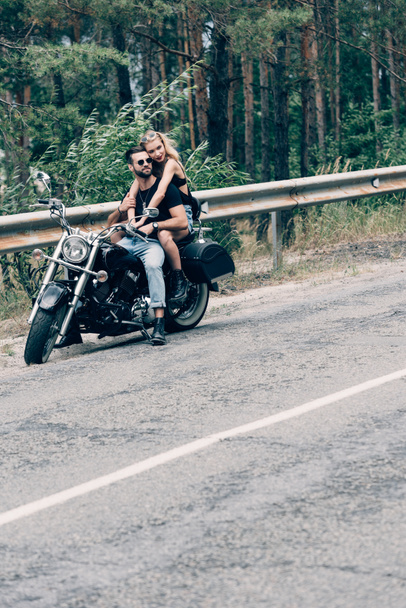 young couple of bikers hugging on black motorcycle on road near green forest - Photo, Image