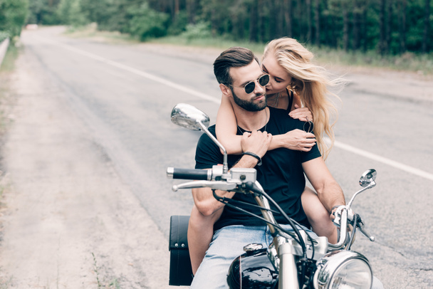 young tender couple of bikers hugging on black motorcycle on road near green forest - Photo, Image