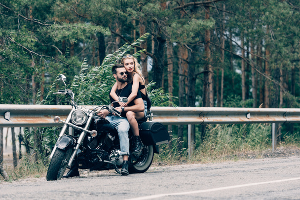 joven pareja de ciclistas abrazándose mientras están sentados en motocicleta negra en la carretera cerca de bosque verde
 - Foto, Imagen