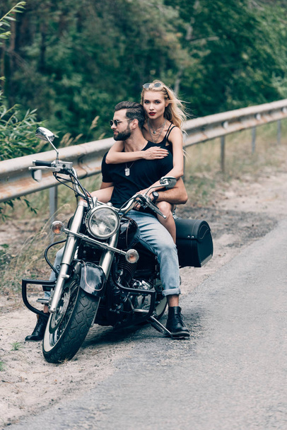 young couple of brutal bikers hugging on black motorcycle on road near green forest - Fotografie, Obrázek